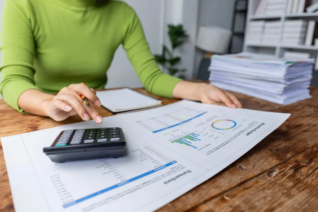 Hands using a calculator while reviewing printed financial charts and reports on a wooden table.