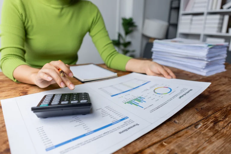 Hands using a calculator while reviewing printed financial charts and reports on a wooden table.