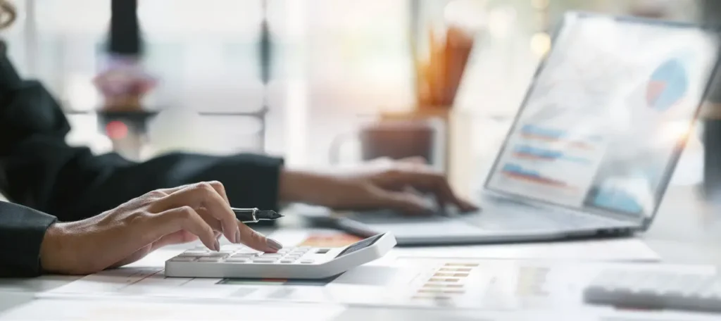 Woman at desk working on calculator with laptop in background.