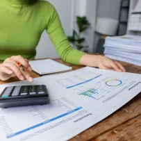 Hands using a calculator while reviewing printed financial charts and reports on a wooden table.
