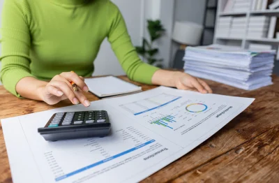 Hands using a calculator while reviewing printed financial charts and reports on a wooden table.
